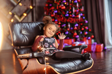 Beautiful kid girl 5-6 year old wearing stylish dress sitting in armchair over Christmas tree in room. Looking at camera. Holiday season.