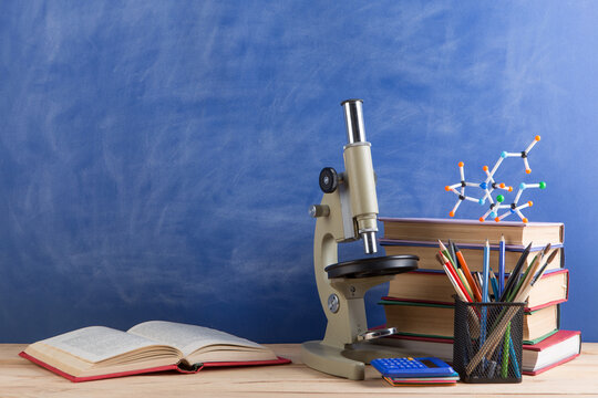 Education and sciences concept - books, molecule model and microscope on the desk in the auditorium, chalkboard background.