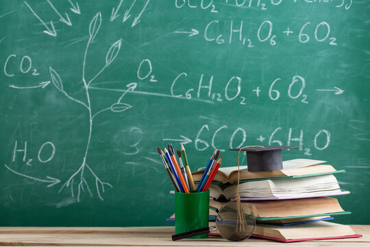 Education and sciences concept - books and graduation cap on the desk in the auditorium, photosynthesis formulas on the background.