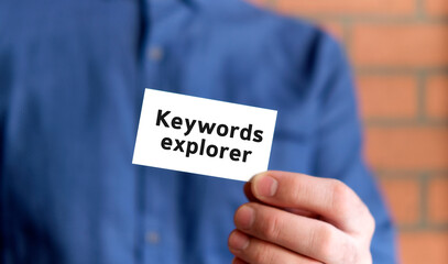 A man in a blue shirt holds a sign with the text of keywords explorer in one hand