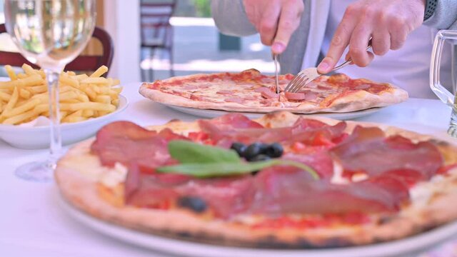 A man cuts pizza at a table in an Italian restaurant. Traditional thin pizza of northern Italy. One pizza is in focus, the other has no focus. Pizza with prosciutto, mazarella and basil.