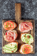 Assorted Italian bruschette snack on a wooden chopping board on a dark textured table. Mini sandwich mix. Top view. Symmetry, close up