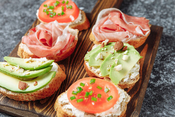 Assorted Italian bruschette snack on a wooden chopping board on a dark textured table. Mini sandwich mix. Close up