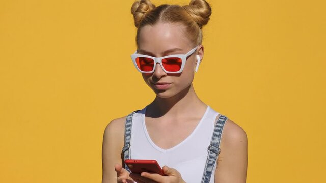 Portrait of happy teen blonde girl in airpods in pink glasses listens to music on smartphone, smiles, dances, shaking her head rhythmically against yellow background of slow motion in summer. Emotions