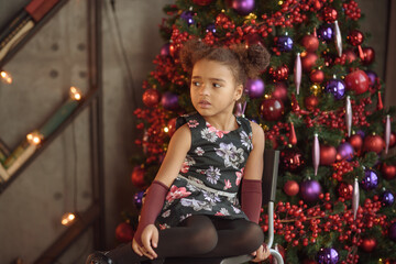 Beautiful kid girl 5-6 year old wearing stylish dress sitting in armchair over Christmas tree in room. Looking at camera. Holiday season.
