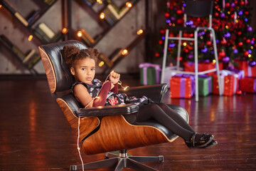 Fototapeta na wymiar Beautiful kid girl 5-6 year old wearing stylish dress sitting in armchair over Christmas tree in room. Looking at camera. Holiday season.