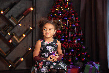 Beautiful kid girl 5-6 year old wearing stylish dress sitting in armchair over Christmas tree in room. Looking at camera. Holiday season.