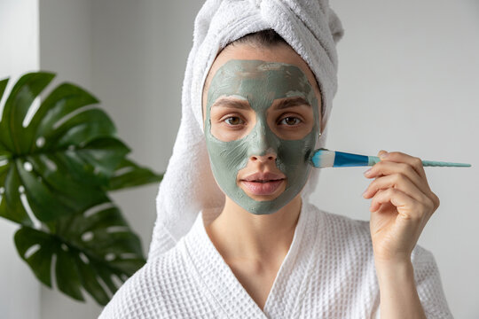 Young woman wearing towel on head and bathrobe putting clay mask on face with brush.
