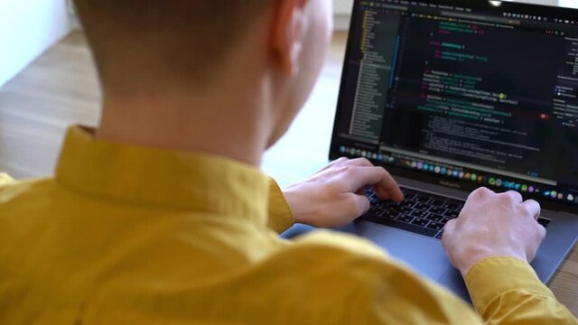 Young programmer businessman in a yellow shirt sitting at a wooden table and typing code on the keyboard, taken from over his shoulder