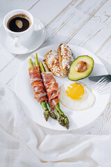 fried asparagus wrapped in jamon, fried eggs, bruschetta with soft cheese and avocado on a white plate and a cup of freshly brewed coffee