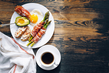 fried asparagus wrapped in jamon, fried eggs, bruschetta with soft cheese and avocado on a white plate and a cup of freshly brewed coffee