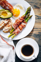 fried asparagus wrapped in jamon, fried eggs, bruschetta with soft cheese and avocado on a white plate and a cup of freshly brewed coffee