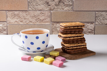 Two biscuits spread on top of chocolatiers with layers of chocolate and milk cream lying on a wooden plank. Nearby is a cup of green tea on a white saucer. Multi-colored sweets in the foreground.