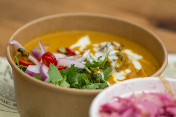 Top view of an Indian yellow curry dish, with chunks of chicken meat, spicy red peppers, yogurt, red onions and green coriander leaves, served in a paper bowl. 