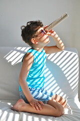 A cute little boy sits in a room and looks through a paper spyglass.