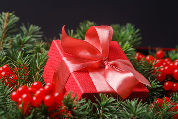 New Year Christmas Xmas holiday celebration red present gift box with satin pink bow, immersed in the needles of a Christmas tree decorated with red berries. Dark festive composition.