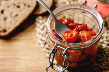 Sun-dried cherry tomatoes olive oil in glass jar with fork, standing on wooden kitchen table with sliced rye bread.