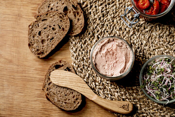 Glass jar of homemade chicken liver pate with wooden knife, sliced rye bread, sun-dried tomatoes and green sprout salad on wooden background. Home breakfast or appetizer. Flat lay