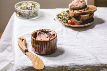 Ceramic bowl of homemade chicken liver pate with wooden knife, sliced rye bread, sun-dried tomatoes and green sprout salad on white linen table cloth. Home breakfast or appetizer
