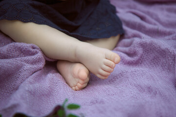 legs peek out from under the dark blue dress of a little girl who sits on a knitted lilac plaid.
selective focus on the toes of the right foot