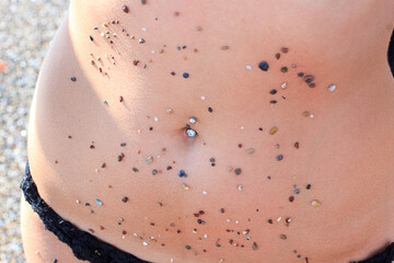 sea stones stuck to the tanned girl’s belly, navel piercing, sunlight falls on the side of the...