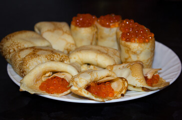 fresh pancakes rolled up in rolls with fish caviar lie on a white plate on a black table.
lmage with selective focus