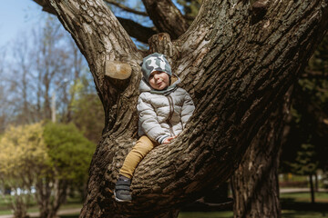 A cute little boy in a camouflage beanie is sitting on the branch of big tree. He is happy to be so high. Image with selective focus