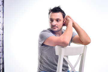 Young man dressed casual, sitting on a white chair, doing face expressions, isolated in white.