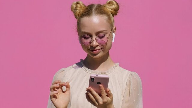 Portrait of happy teen blonde girl in airpods in pink glasses listens to music on smartphone, smiles, dances, shaking her head rhythmically against pink background of slow motion in summer. Emotions