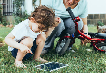 A young boy with his mother sitting on the grass in a park using a tablet PC