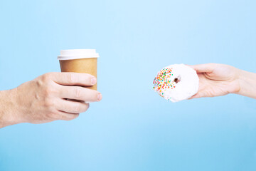 Man hand holds paper cup with coffee, woman hand holds donut with white icing and multicolored sugar pastry topping, blue background