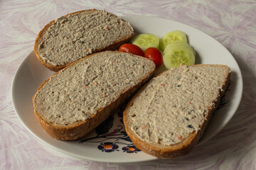 A modest breakfast in the form of three breads with fish spread and as a side dish are small tomatoes and sliced cucumber. Everything ready on a plate to eat.