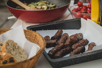 Home made cevapcici meal on a picnic. Cevapcici in a metal container with paper towel, served surrounded with bread basket and different salads.