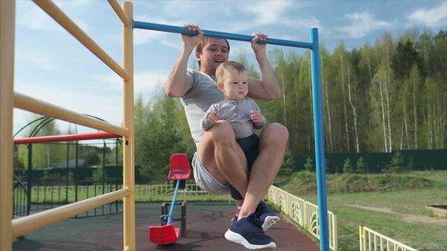 Family work out. Young man does pull ups with his baby. Adult male performs work out with his little child as additional weight.