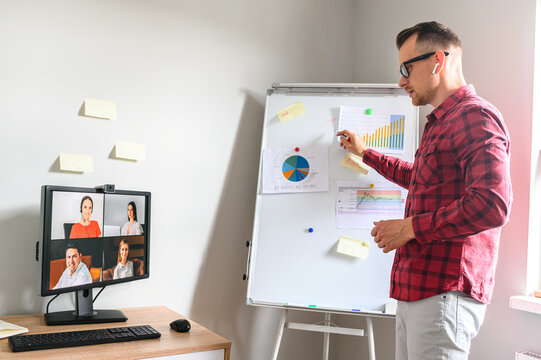 Confident young man perform a presentation online to his clients using a flipchart via PC app for video conferences, he stands in the office and points on a flipchart