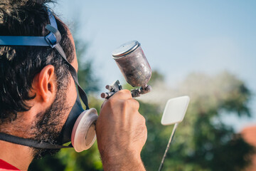 Amateur painter in red sweater and face mask using a spray gun to paint a plastic object or part in a home garden. Back view of a painter with a small spray gun. Focus on the gun