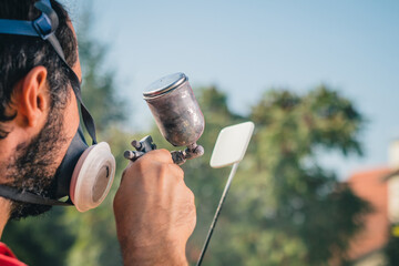 Amateur painter in red sweater and face mask using a spray gun to paint a plastic object or part in a home garden. Back view of a painter with a small spray gun. Focus on the gun