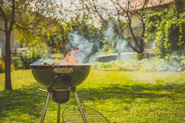 Round mobile grill or barbecue parked on a green surface or lawn. Visible flame and fire on a wooden and coal fuel with thick smoke rising up from the grill.