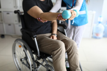 Man on wheelchair works out his hand with dumbbell. Simple exercises for disabled. Doctor helps...