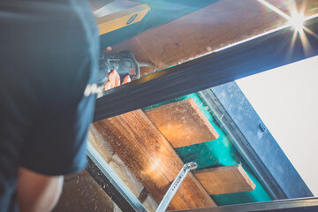 A person is cutting a board of wood next to a window with a reciprocating saw. Sawdust is flying around.