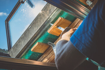 A person is cutting a board of wood next to a window with a reciprocating saw. Sawdust is flying...