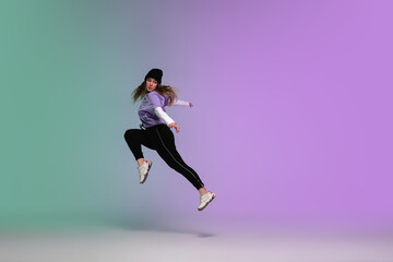 In jump. Beautiful girl dancing hip-hop in stylish clothes on colorful gradient background at dance hall in neon light. Youth culture, movement, style and fashion, action. Fashionable bright portrait.