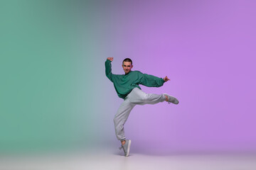 Beautiful sportive boy dancing hip-hop in stylish clothes on colorful gradient background at dance...