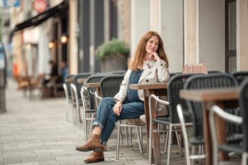 Beautiful young pregnant woman wearing sweater, jeans, grey coat sitting outside a cafe with a beautiful exterior in the summer.