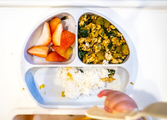 Combination menu for baby, white rice, pumpkin fried with egg and basil, piece of sliced of strawberries in funny silicone dish on white table