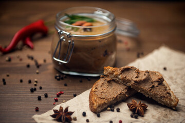 Poultry pate spread on bread and glass jar with poultry pate, decorated with almonds and rosemary, and a few fruits of hot pepper on a wooden table