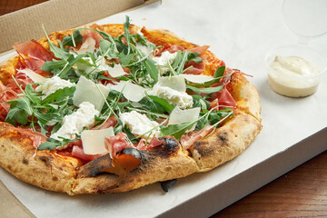 Photo for pizza delivery. Pizza with jamon, parmesan, arugula and strachatella cheese in a cardboard box with side sauce on a wooden table