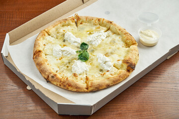 Photo for pizza delivery. 4 cheese pizza with blue cheese, mozzarella, parmesan and straccella in a cardboard box with side sauce on a wooden table