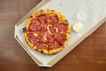 Photo for pizza delivery. Pizza with spicy chorizo salami, melted cheese in a cardboard box with side sauce on a wooden table. Close up