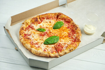 Photo for pizza delivery. Pizza margherita with tomatoes, basil and mozzarella in a cardboard box with sauce for sides on a wooden table